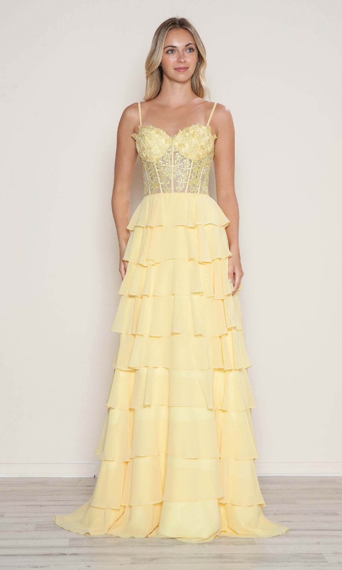 A woman stands against a plain wall wearing the Poly USA 9698 Long Tiered Corset Prom Dress in pastel yellow, featuring spaghetti straps and a flowing ruffle A-line skirt.